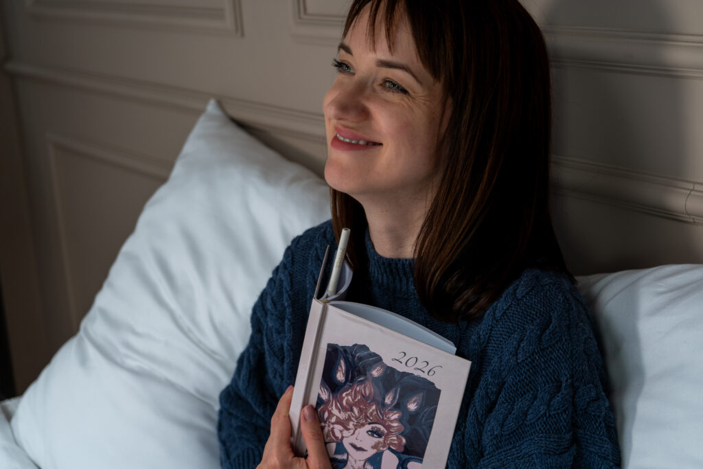 Kobieta trzyma kalendarz THIStanS 2026 z uśmiechem, siedząc w łóżku w przytulnym swetrze - spokojne planowanie roku bez presji